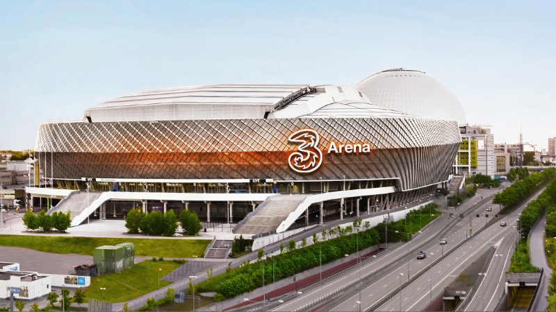 3Arena i Stockholm, en modern byggnad med en glänsande, silvergrå fasad i ett flätat nätmönster. Mitt på fasaden sitter en stor, lysande logotyp för telekombolaget 3. Arenan har breda trappor och ingångar i flera plan. I bakgrunden till höger skymtar den vita kupolen av Avicii Arena (tidigare Globen) mot en ljusblå himmel. Arenan är fotograferad från en upphöjd vinkel och ramas in av grönska samt en flerfilig bilväg i förgrunden.