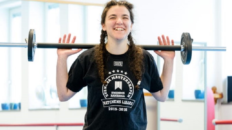 En ung kvinna med brunt hår i flätor och en svart t-shirt håller en skivstång på axlarna och ler stort.
