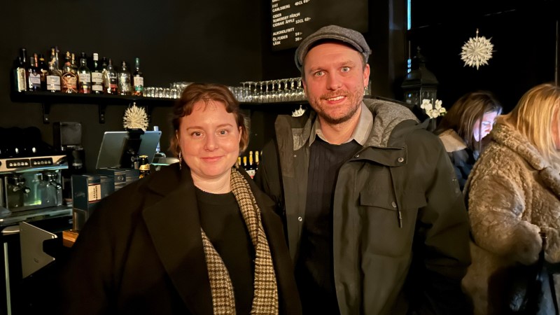 En vit man och kvinna i 35-årsåldern står framför en bardisk. Rummet är svartmålat. De har ytterkläder på sig.