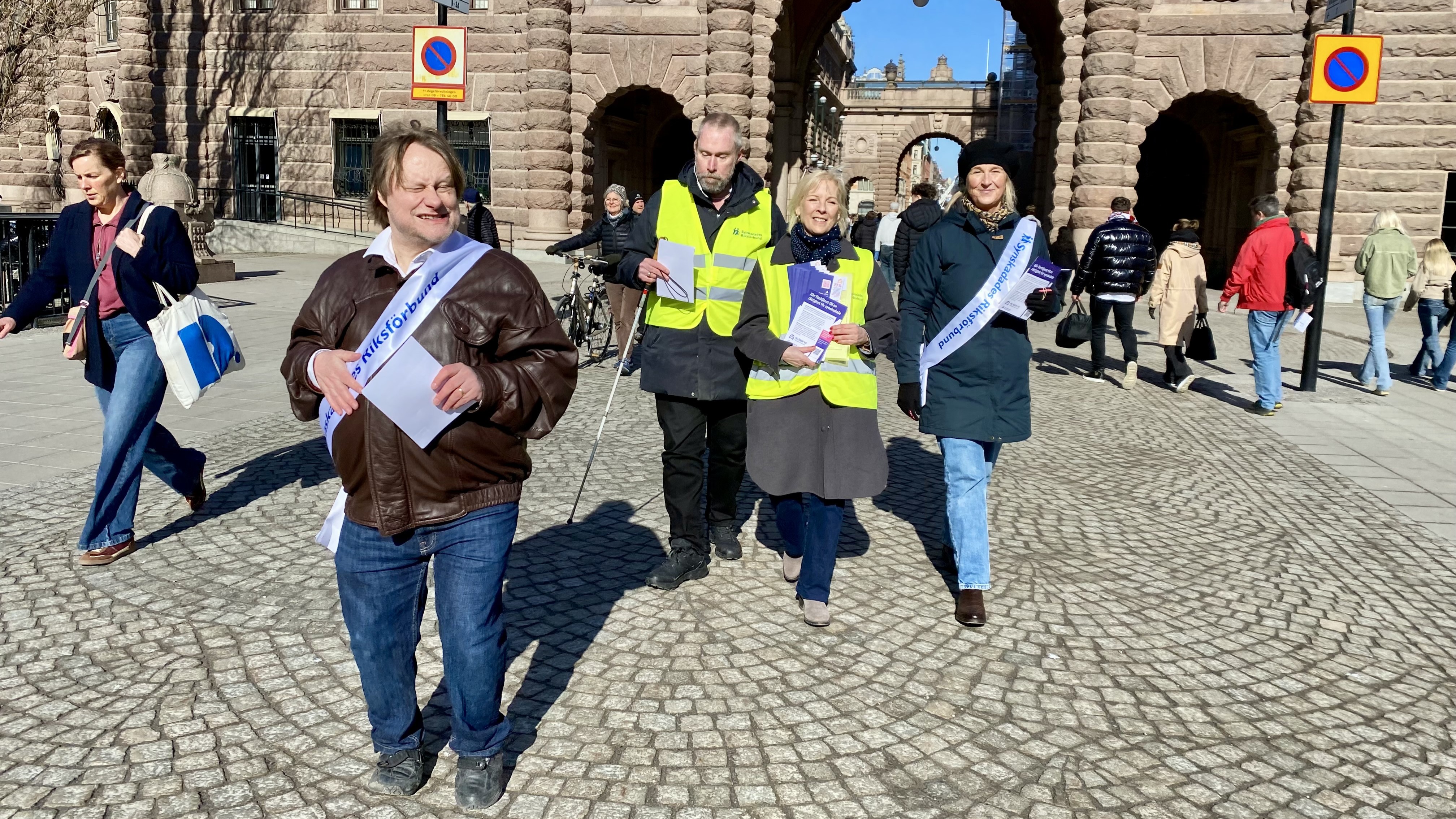 På Mynttorget vid riksdagshuset, går några personer med SRF:s ordensband i vitt och har broschyrer som de delar ut till förbipasserande.