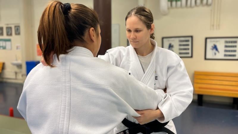 Två vita kvinnor i 30 års-ålders greppar varandra i judo. De har vita judodräkter och är i en träningshall.
