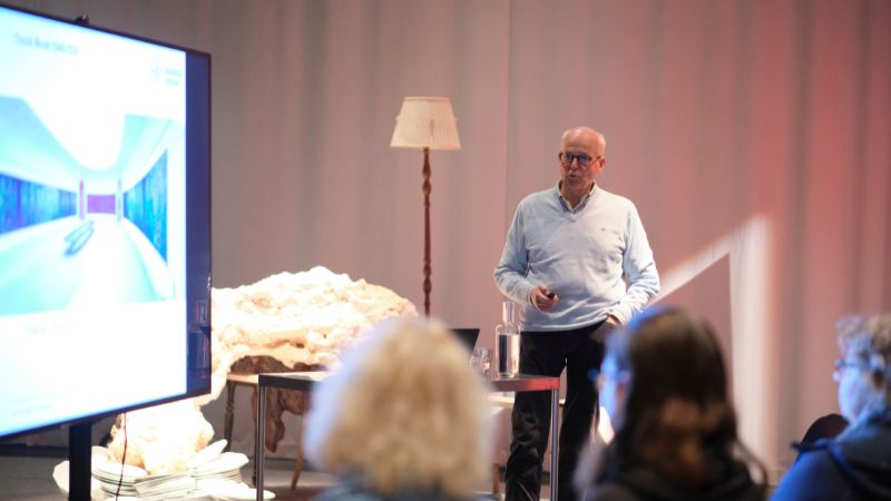 Äldre vit skallig man med glasögon, håller föredrag framför publik. En stor TV-skärm i rummet.