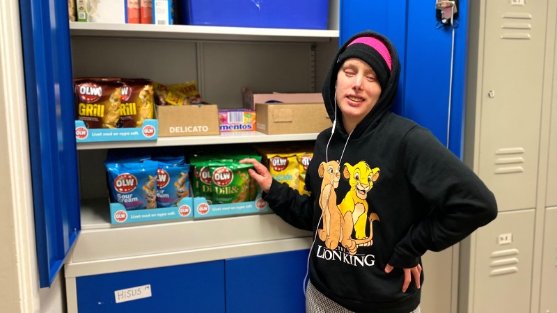 Ung rödlätt kvinna, i svart hoodie. Står vid ett öppet blått skåp fyllt med chips och godis