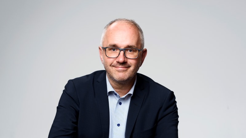 Man i blå skjorta och svart kavaj. Han har rektangulära glasögon och kort tunnt grått hår.