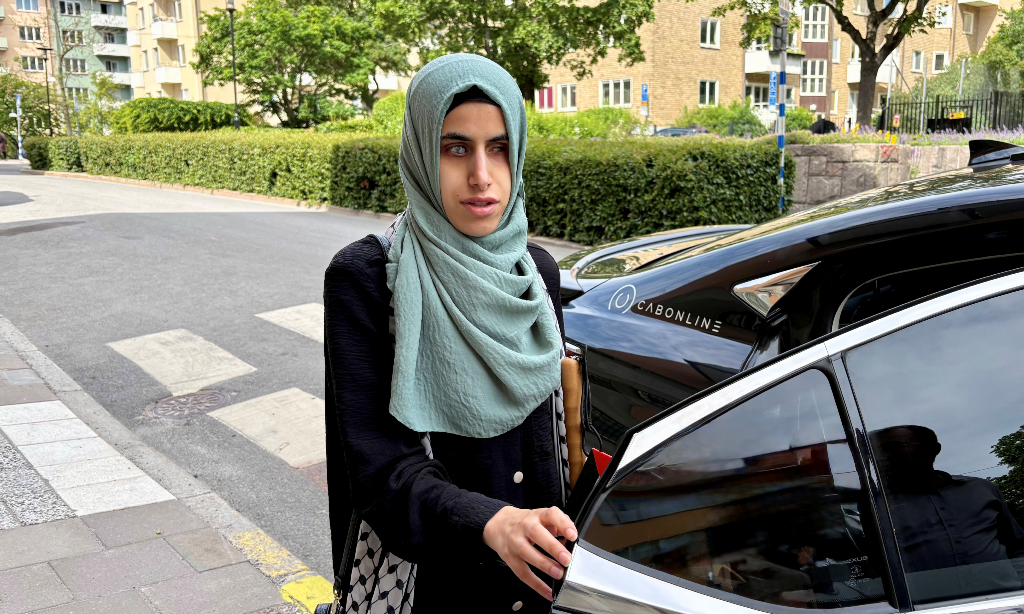 En ung kvinna med ljus hy och muslimsk grågrön schal runt huvudet, står på gata på väg in i en färdtjänsttaxi. Hon har svart lång klänning och vit käpp.