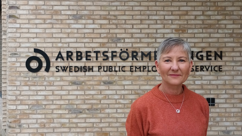 En kortklippt gråhårig kvinna framför en gul tegelfasad med texten Arbetsförmedlingen på väggen.