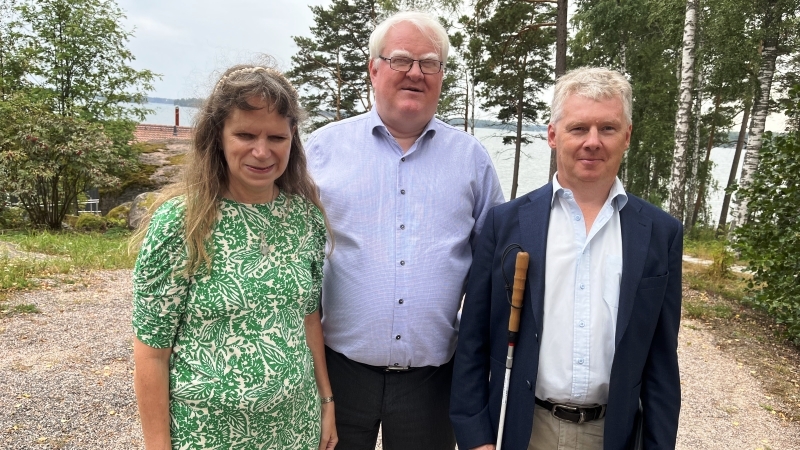 Tiina Nummi Södergren har en grön- och vitblommig klänning och långt vågigt hår. Terje Andre Olsen har vitt hår, glasögon och en ljusblå skjorta. Niklas Mattsson har ljusgrått lockigt hår, ljus skjorta och mörk kavaj. Han håller i en vit käpp. De står i en skogsbacke med en havsvik bakom sig