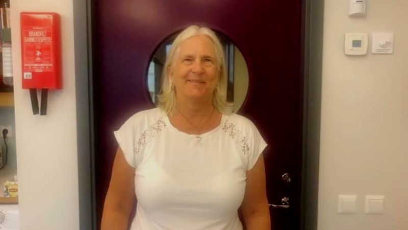 Bild i halvfigur på småleende kvinna med blond axellångt hår i mittbena, klädd i vit t-shirt. Hon står framför en mörk dörr med ett runt fönster i huvudhöjd.