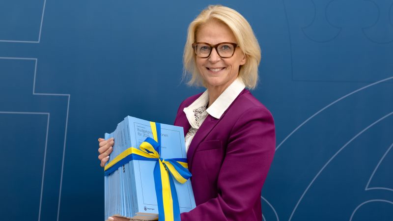 Blond leende vit 60-årig kvinna i page. Lilarosa kavaj, vit spetsblus under. Håller i en rejäl bunt med ljusblåa häften. De har ett blågult sidenband runt sig med rosett.