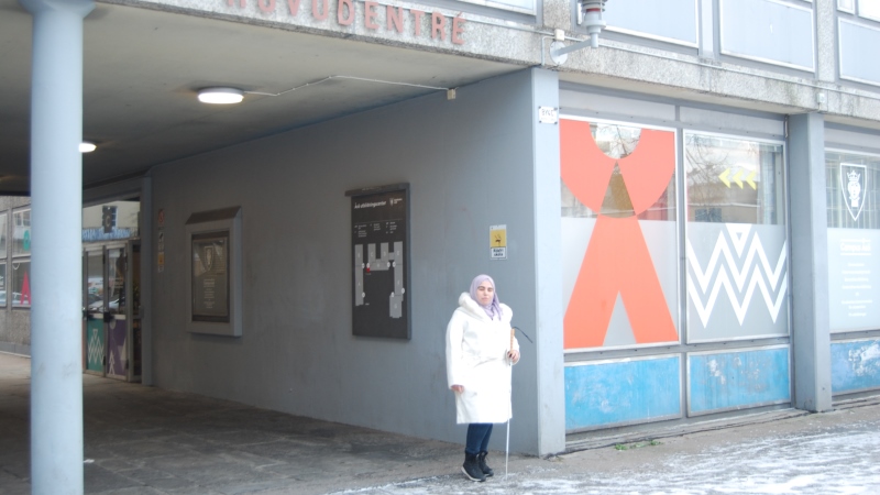 Vid en gångtunnel står en kvinna i vit kappa och huvudduk, vit käpp i handen.