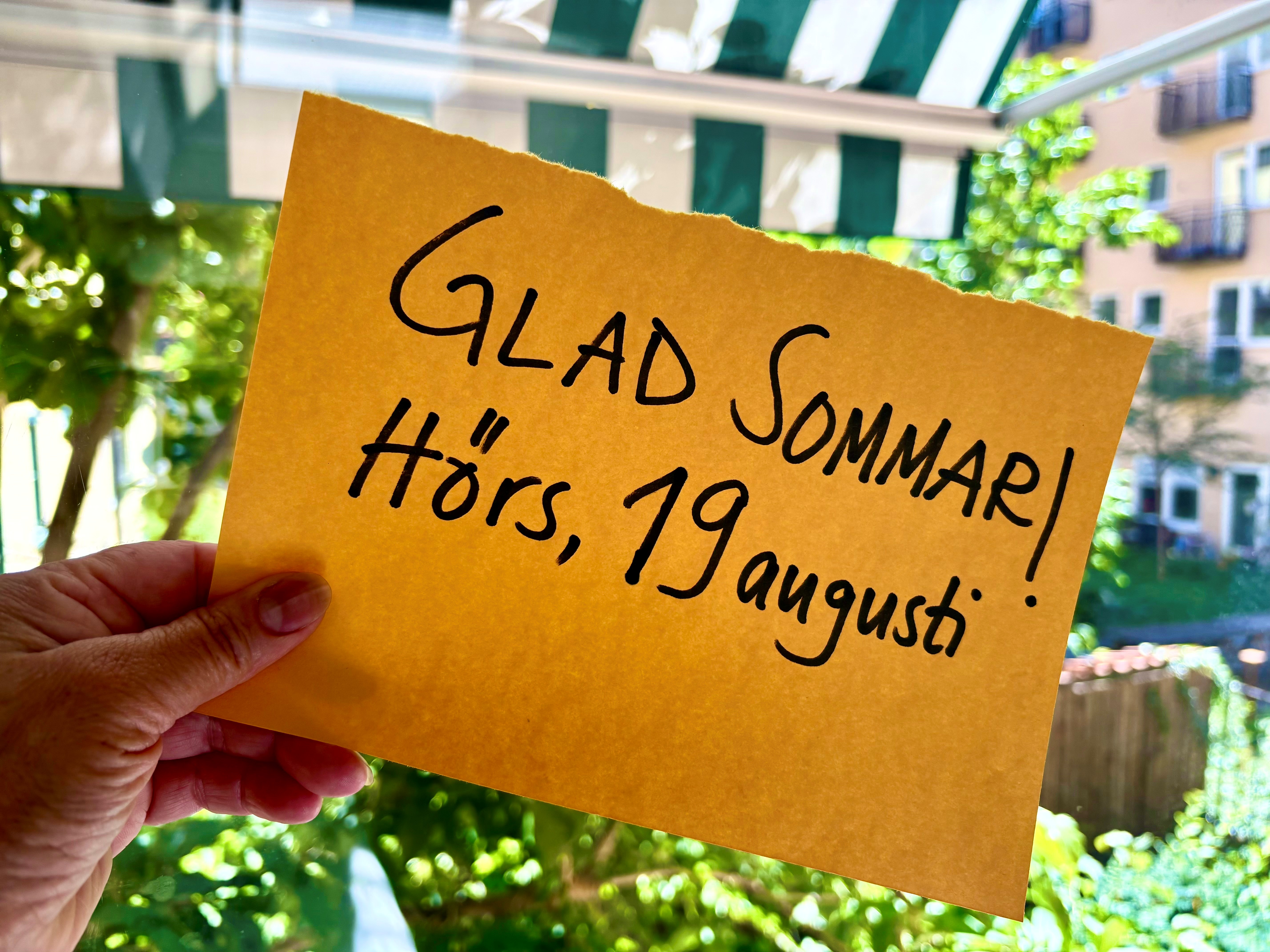 En hand håller upp en gul lapp framför ett fönster med grön utsikt. På lappen står: Glad sommar! Hörs, 19 augusti