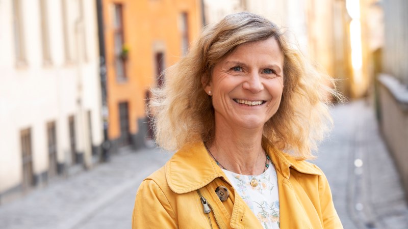 Leende kvinna med axellångt vågigt ljus hår, gul jacka, står i stadsmiljö