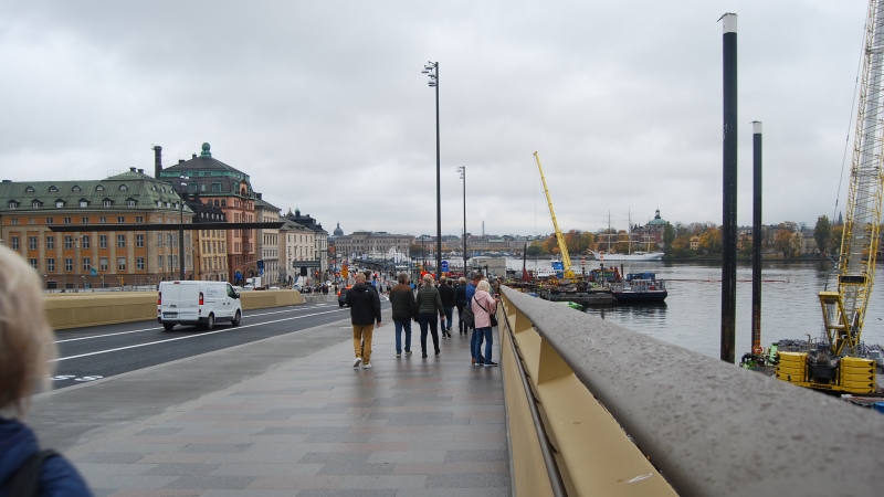 Gående människor och bilar på bron i riktning mot Gamla stan, inramade av de guldkantade räckena i varm, lätt skimrande guldfärg.  