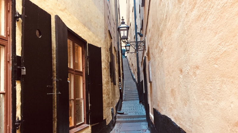En mycket smal gatugränd med en lång uppåtgående trappa mellan husväggarna. Dessa är i ljusgul puts. Fönsterluckor i trä till vänster. Bildtext: Mortens Trotzigs gränd i Gamla stan.