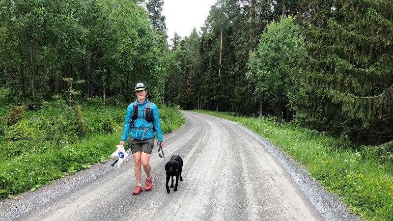 Yngre, vit kvinna går på grusväg i skog, med svart ledarhund. Hon har  grågröna shorts, röda jympaskor, turkos jacka och keps. Under kepsen har hon pannbandet med sändaren. Hon har hörlurar på sig.