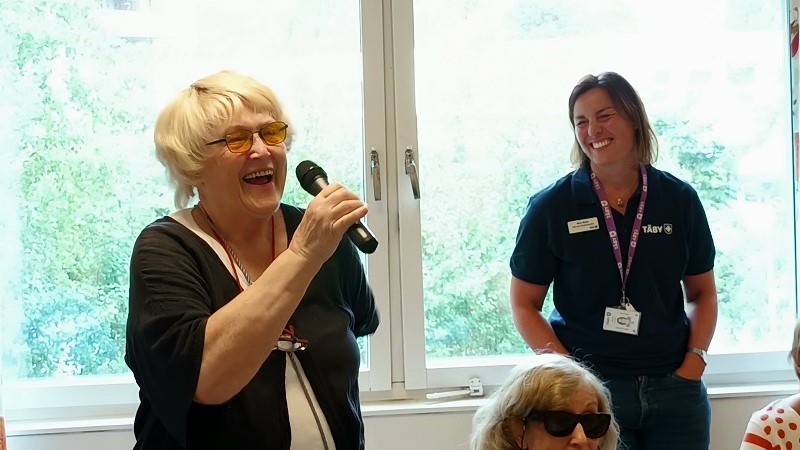 En blond äldre kvinna  (Maritta Söderholm) håller en mikrofon och skrattar, till höger om henne står en yngre kvinna (Nina Stehn) med brunt axellångt hår och ler stort. Det står Täby på bröstet på hennes mörkblå arbetströja. I bakgrunden ett fönster med grönska utanför.