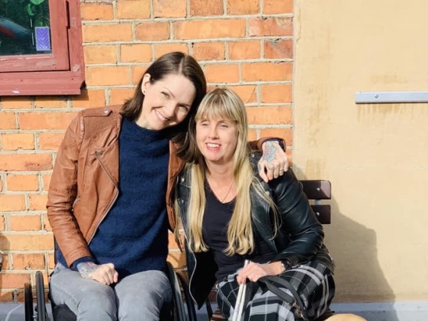 Kvinna med ljust hår och vit käpp sitter bredvid brunhårig kvinna i rullstol vid en husvägg
