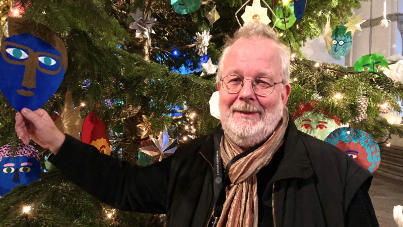 En tunnhårig vithårig rundlagd man med skägg står vid en stor julgran, han håller i ett julpynt i form av ett blått ansikte.