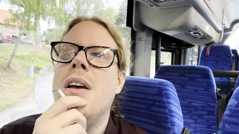 En vit man i 40-årlsåldern sitter på en buss med blå säten, bär glasögon och håller en vit sladd med mikrofon vid munnen. Han har blont långt hår uppsatt i en hästsvans. Bussens tomma säten och träd syns i bakgrunden.