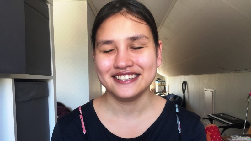 Porträttbild på en brett leende tonårstjej med mörkt hår i mittbena samlat i tofs i nacken. Över de slutna ögonen har hon tjocka mörka ögonbryn och är klädd i en svart tröja.