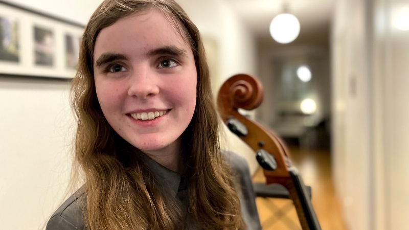 Ung vit leende tjej, brunt axellångt hår. Står i korridor och håller i en cello.