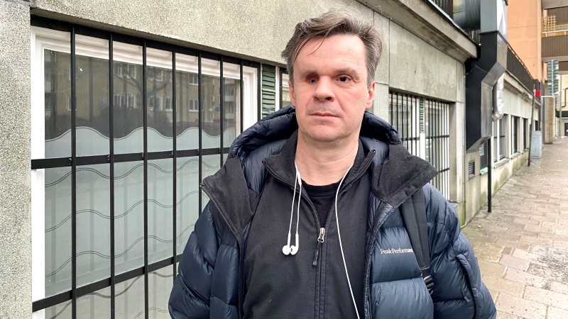 Vit medelålders man, brunt kort hår, mörkblå täckjacka. Svart hoodie under. Står på trottoar, i gråmulen stad.