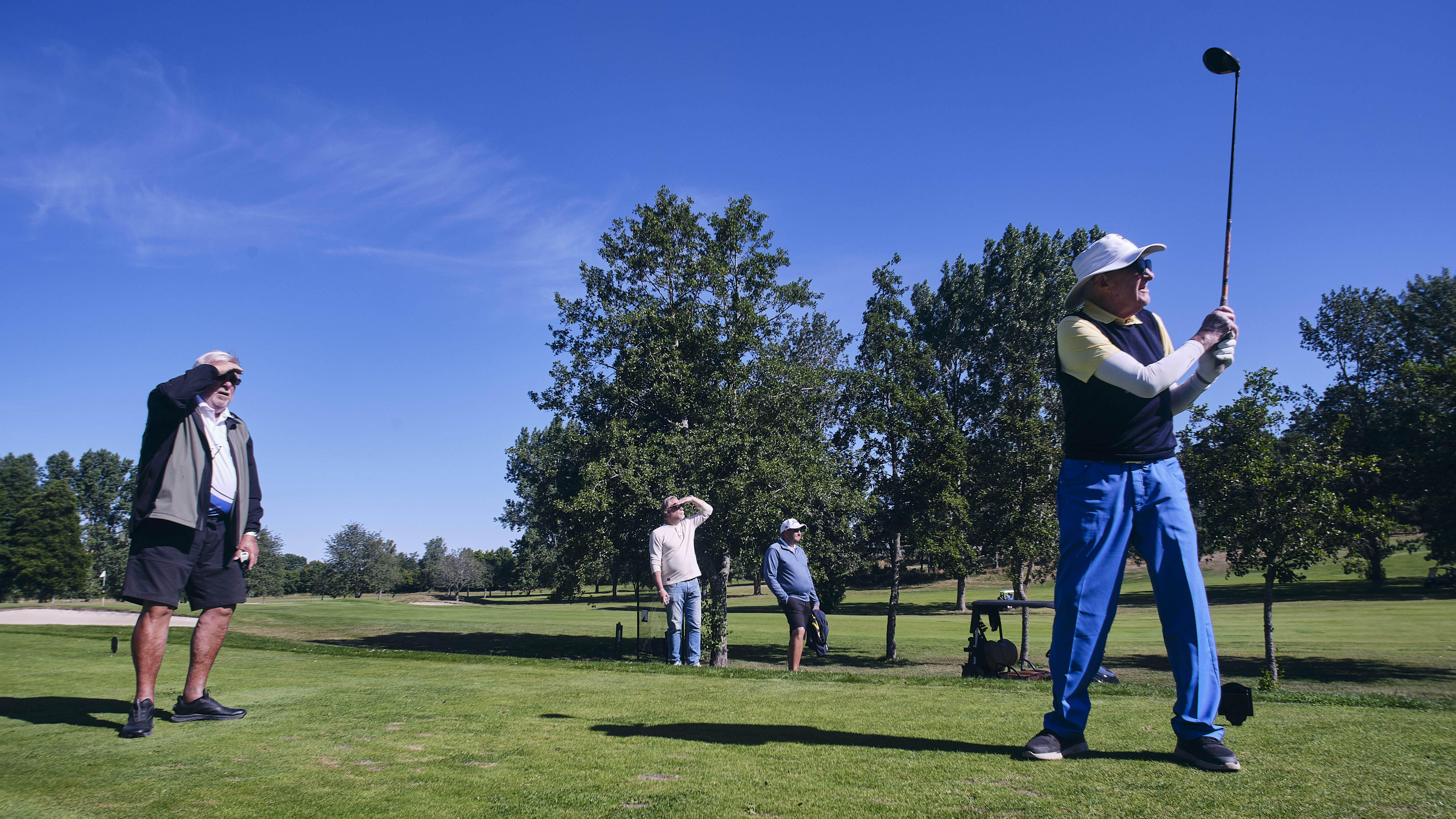 En äldre man i solhatt står med golfklubban höjd efter ett slag, en bit bort bakom honom tre andra män som spanar efter bollen utanför bild. Golfbana med träd i bakgrunden.