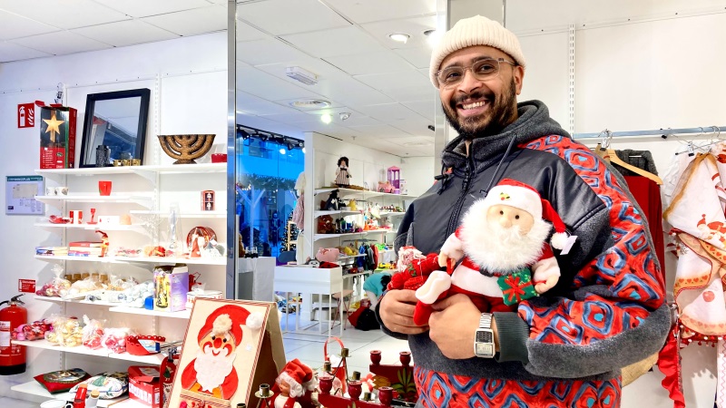 En mörkhyad medelålders man med skägg och glasögon håller i två mjukistomtar. Står i secondhandbutik med julprydnader i hyllorna.