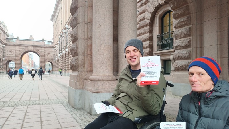Två män, den ena i rullstol, sitter utanför riksdagen med flygblad i handen. 
