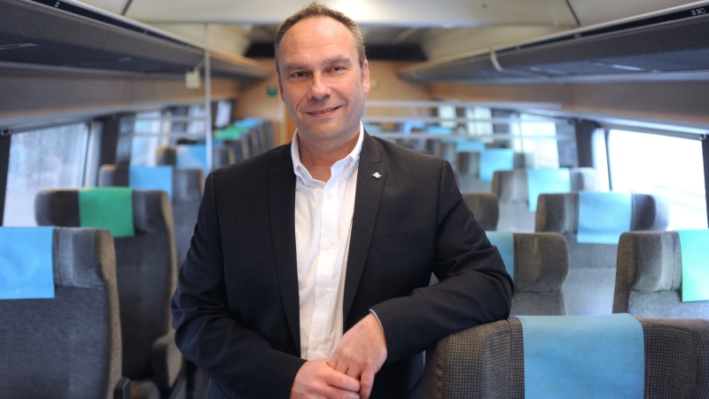 Korthårig man med mörkgrå kavaj och vit skjorta står i en tågvagn med toppa sittplatser.