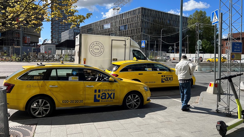 Två gula taxibilar på en gata. En man i vit jacka står på trottoaren. I bakgrunden det ena av Norra Tornen vid Torsgatan, Stockholm. 