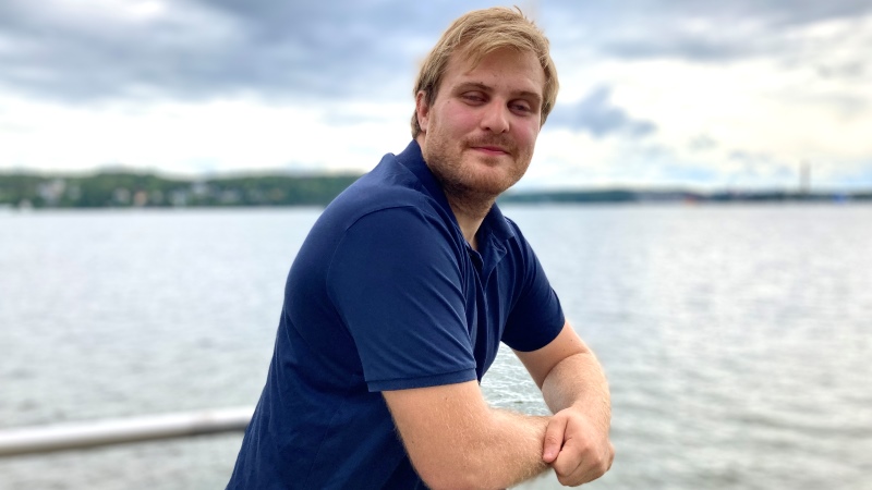 Ung vit man, ljust hår, mörkblå t-tröja, står lutan mot balkongräcke, bakgrund hav