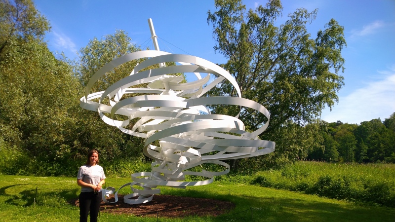 Kvinna långt mörkt hår, grå t-shirt och svarta byxor står i lummig parkmiljö och ser liten ut framför en sju meter hög vit skulptur i form av en tornado..