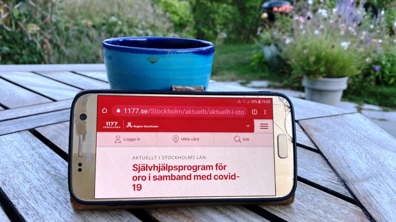 En smart telefon står lutad mot en kaffekopp på ett trädgårdsbord. I bakgrunden skymtar en trädgård och skärmen visar 1177.se och texten Självhjälpsprogram för oro i samband med covid-19.