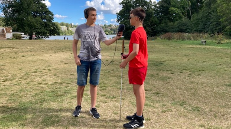 Två tonårskillar i shorts på grässlänt, sommar. Den ena håller i en mikrofon.