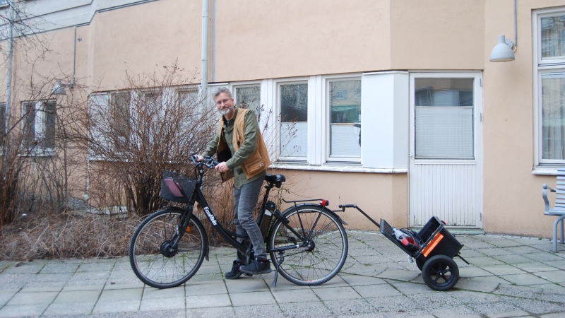 Gråhårig man på cykel med liten kärra bak, han har gul väst, grön tröja. Buskar, hus i bakgrund.