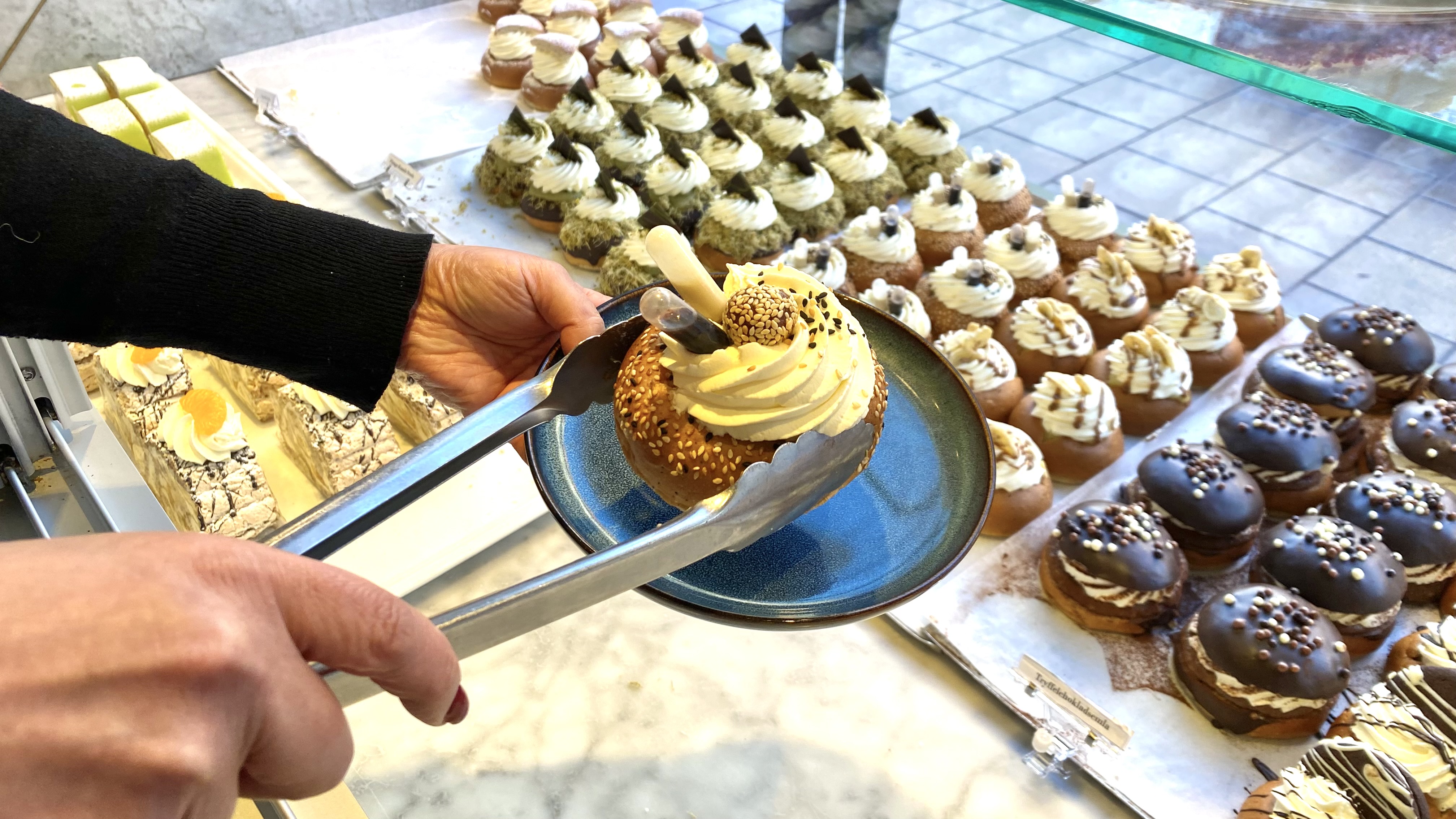 På insidan av skyltfönster på ett bageri, ser man flera semlor av olika slag. Händer syns i förgrunden, den ena håller i en talrik den andra i en tång med en semla ovanför tallriken.