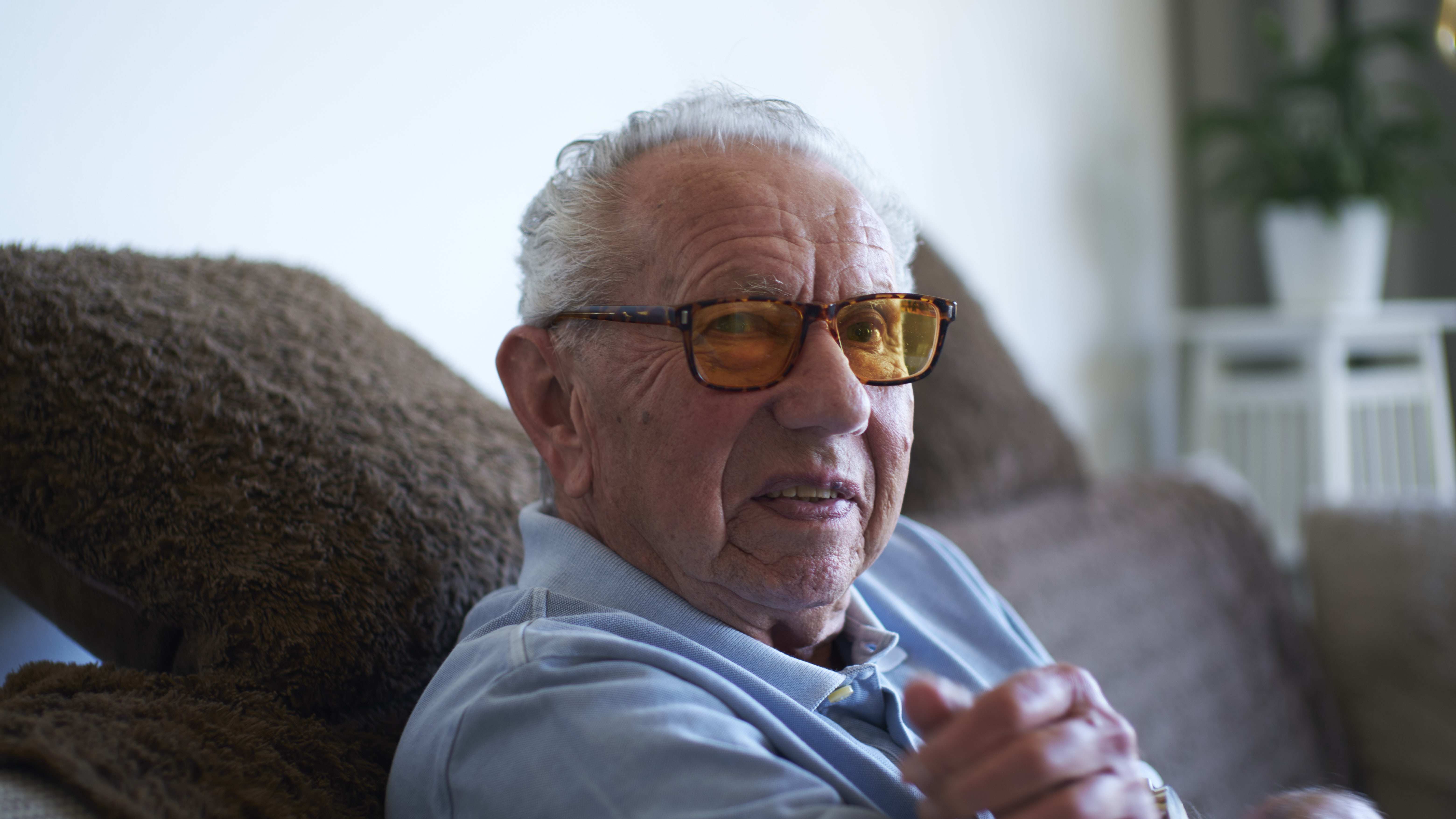 Roger Lindroth, en 79-årig vit man sitter i en brun soffa. Han har kort hår kammat bakåt, bär en ljusblå skjorta och rektangulära glasögon med gult glas och spräckliga bågar.