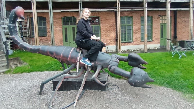 En ung kvinna med långt blont hår sitter på ryggen av en jättestor modell av en skorpion framför en tegelbyggnad.