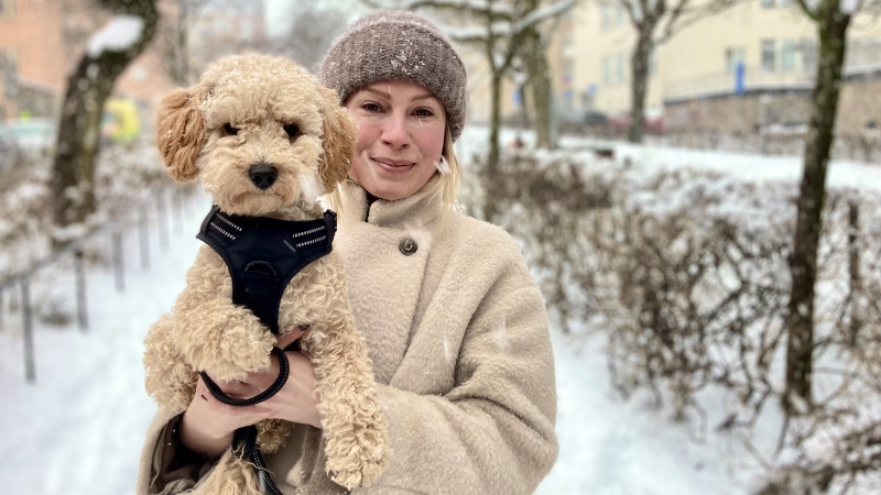 En blond, ljus kvinna står med sin lilla pudel i famnen, i en snöig trädallé.