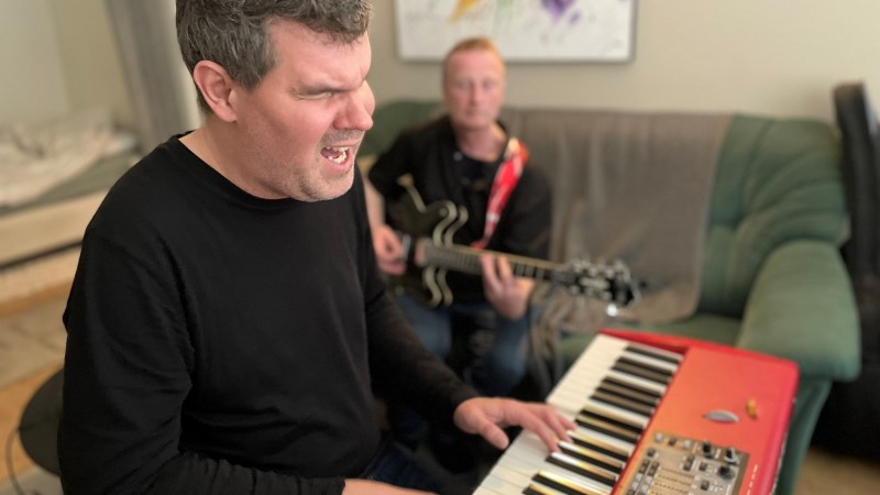 En vit medelålders man med kort brunt hår och svart tröja, sitter och spelar keyboard och sjunger, vid sidan om honom i en soffa, sitter en vit något äldre man och spelar elgitarr. De är i ett vardagsrum. 