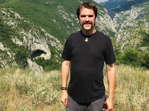 40-årig vit man med vågigt brunt kort hår, och mustasch, svart t-shirt. Står i bergslandskap. Sommar.