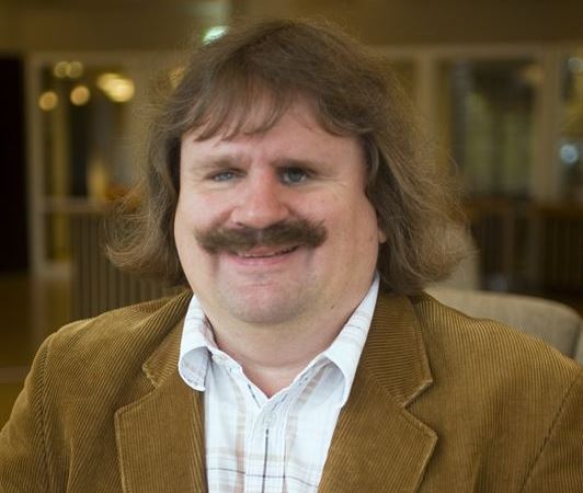 Vit medelålders man, med kraftig brun mustasch. Ljusbrun kavaj och vit skjorta.