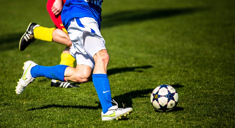 Bild på två springande fotbollsspelares ben. Den ena har blåa strumpor den andra har gula. Vit fotboll och grön gräsplan. Bild på två springande fotbollsspelares ben. Den ena har blåa strumpor den andra har gula. Vit fotboll och grön gräsplan.