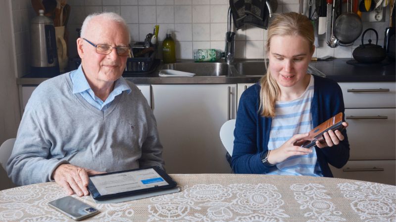 En äldre vit man med glasögon med blå skjorta under en grå pullover sitter vid ett köksbord. Framför honom ligger en Ipad och en mobiltelefon. Till höger om honom sitter en vit kvinna i 25-årsåldern med långt blont hår i en hästsvans. Hon har en vit-blå-randig tröja med en blå kofta över. I Handen har hon en mobiltelefon. De sitter i köket.