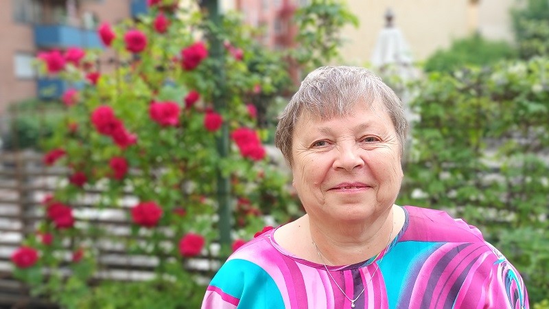 En gråhårig kvinna med skrattrynkor och en flerfärgad klänning i lila och rosa, i bakgrunden en rosbuske med många blommor.