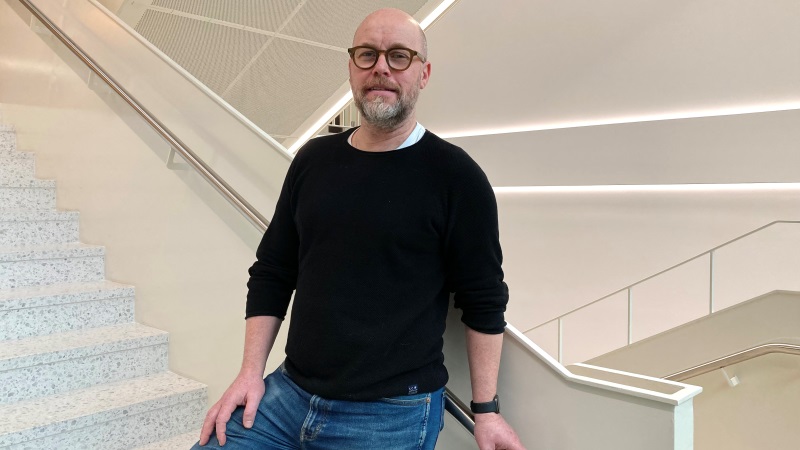 Vit medelåldersman, med stubbat hår och grått skägg. Svart långärmad tröja, blå jeans. Glasögon. Står i vått trapphus.