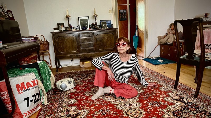 En 80-årig smal kvinna med rödbrun page och mörka glasögon, sitter på golvet i sitt vardagsrum. Röd Persisk matta. Hon har en svartvit randig tröja och röda byxor.