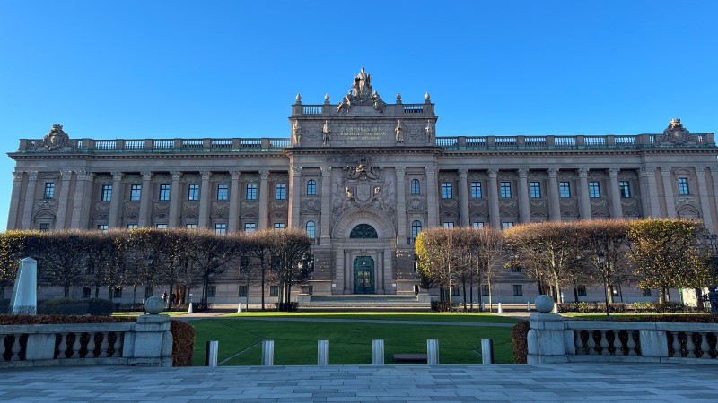 Riksdagshuset från Riksplan. Stor pampig byggnad i varmbeigefärgad sten. Stor öppen plats framför med trädallé.