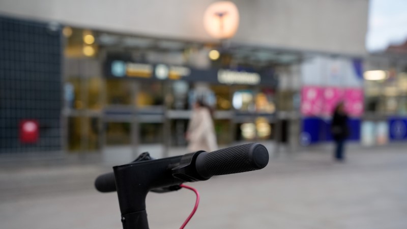 Närbild av styret på en elsparkcykel, bakgrund: ingång till tunnelbanestation